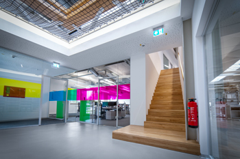 Dein Profil Helles Foyer mit moderner Glaswand in kräftigen Farben, Holztreppe und Zugang zu einem offenen Bürobereich in einem zeitgemäßen Bürogebäude.