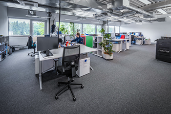 Deine Aufgaben Weiträumiges, modernes Büro mit Einzelarbeitsplätzen, großen Fenstern, Pflanzen und offenem Deckensystem für eine kreative Arbeitsumgebung.